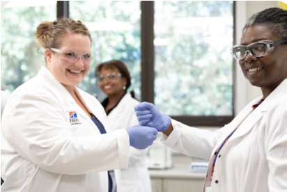 Twee wetenschappers in laboratoriumjassen en handschoenen glimlachen en schudden elkaar de hand in een laboratoriumomgeving.