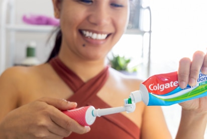 Een vrouw die glimlacht terwijl ze Colgate-tandpasta op een tandenborstel doet in een badkamer.